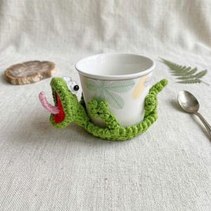 Può includere: Una tazza in ceramica bianca con un motivo a foglie di palma verde chiaro, avvolta da un copritazza all'uncinetto a forma di lucertola verde brillante. La lucertola ha la bocca spalancata con interno rosso, lingua rosa e due occhi bianchi.