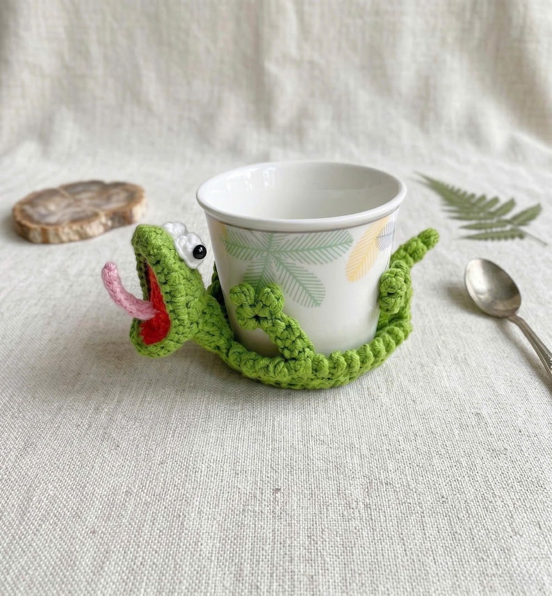 A vibrant green crocheted lizard coaster tightly gripping the base of a white ceramic coffee mug with its four legs. The lizard has a wide-open red mouth cavity, a pink 3D tongue reaching outwards, and two bulging white amigurumi eyes with black safety pupils.