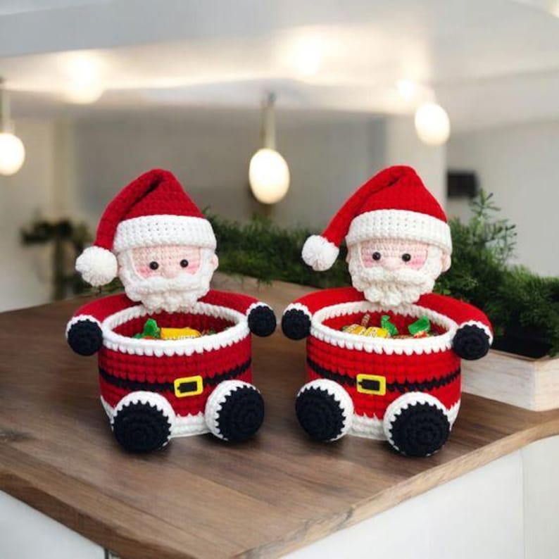 May include: Two crocheted Santa Claus figures with red hats, white beards, and red suits. The Santa figures have black arms and legs, and a yellow belt buckle. The open basket-like bodies are filled with colorful candies. The figures are on a wooden surface.