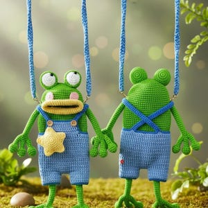 Puede incluir: Dos adornos de rana de ganchillo hechos a mano. Las ranas son verdes con ojos blancos, con petos azules. Una rana tiene una estrella amarilla en su peto. Están suspendidas por cuerdas azules, sobre un fondo verde borroso.