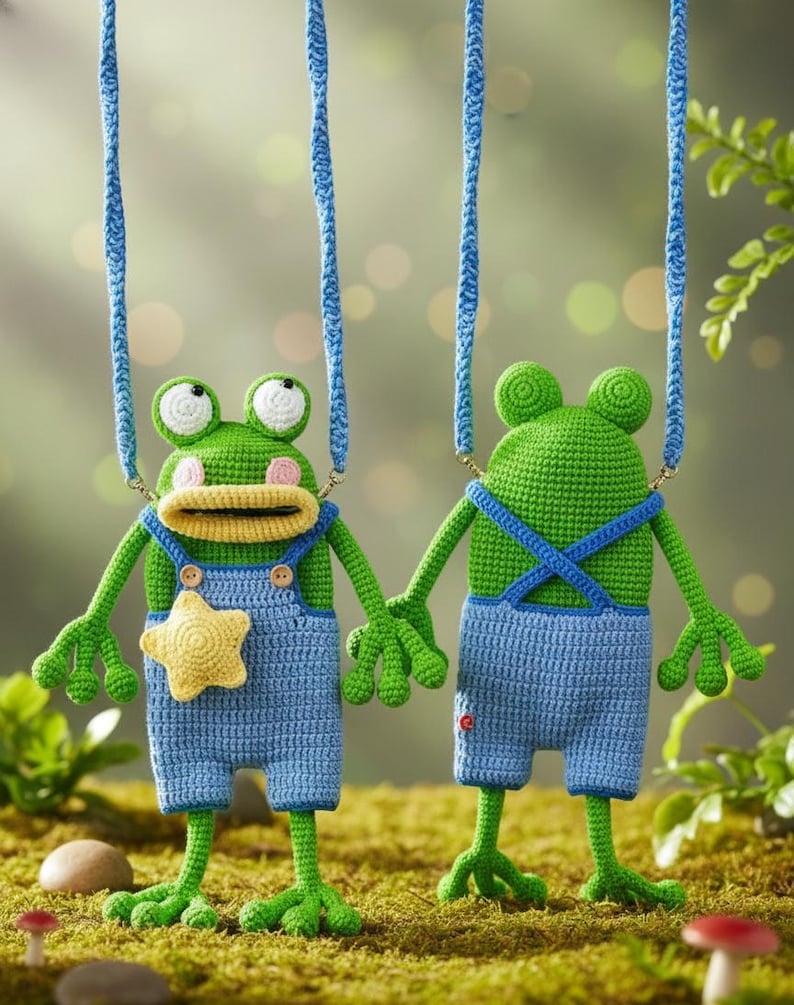 May include: Two handmade crochet frog ornaments. The frogs are green with white eyes, wearing blue overalls. One frog has a yellow star on its overalls. They are suspended by blue cords, set against a blurred green background.