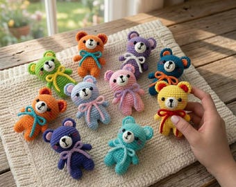 May include: A collection of colorful, handmade crochet teddy bears. Each bear features a unique color scheme, including orange, blue, pink, and yellow, with contrasting bows and embroidered facial details. The bears are arranged on a cream-colored, textured surface.