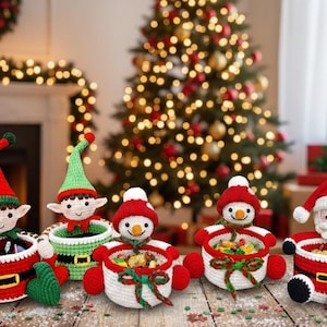 May include: Five handmade crochet Christmas characters are displayed: two elves, two snowmen, and Santa Claus. Each character has a basket body, red and green accents, and is filled with colourful sweets. A decorated Christmas tree is in the background.