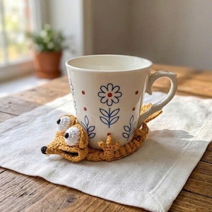 May include: A white ceramic mug with a blue and red floral pattern sits inside a mustard yellow crocheted coaster shaped like a lizard. The coaster features two large white eyes with black pupils and a small black nose, wrapping around the mug base.