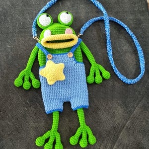 May include: A handmade crochet frog-shaped bag with a blue strap. The frog has green skin, white eyes, and a yellow mouth. It wears blue overalls with a yellow star. The bag is designed to be worn over the shoulder.
