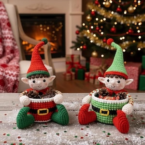 May include: Two crocheted elf figures, one red and one green, each holding a basket filled with pinecones and candies. The elves have pointed hats and are decorated with a black belt and gold buckle. Christmas tree and fireplace in the background.