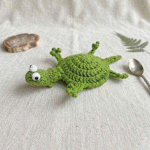 May include: A bright green crocheted turtle toy featuring a round, textured shell, four small legs, and a head with two prominent white and black eyes. The yarn work displays a consistent stitch pattern throughout the entire body of the creature.