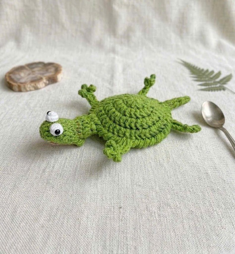 May include: A bright green crocheted turtle toy featuring a round, textured shell, four small legs, and a head with two prominent white and black eyes. The yarn work displays a consistent stitch pattern throughout the entire body of the creature.