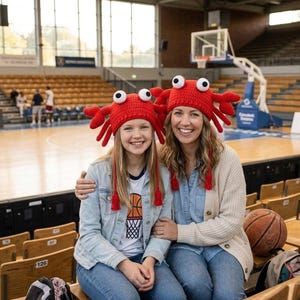 May include: Two people wearing matching red crab-shaped crochet hats with googly eyes and dangling tassels. They are sitting in a basketball arena. The hats have crab claws and are a bright red colour.