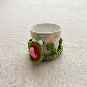 May include: A white ceramic cup featuring a palm leaf pattern, wrapped in a green crocheted frog cozy. The frog has a wide open mouth with a red interior and a small pink tongue, with two small arms extending around the sides of the cup.