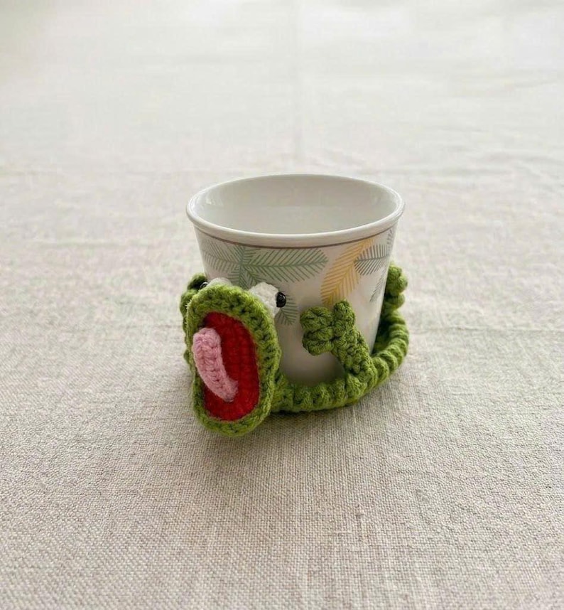 May include: A white ceramic cup featuring a palm leaf pattern, wrapped in a green crocheted frog cozy. The frog has a wide open mouth with a red interior and a small pink tongue, with two small arms extending around the sides of the cup.
