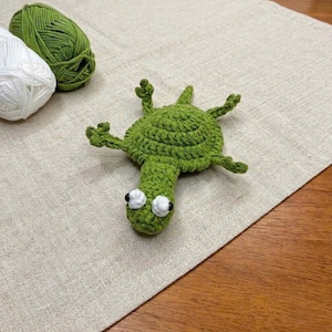 May include: A green crocheted turtle figure with a round shell, four small legs, and a head featuring two white eyes with black pupils. The turtle is positioned near a ball of green yarn and a ball of white yarn on a textured fabric surface.