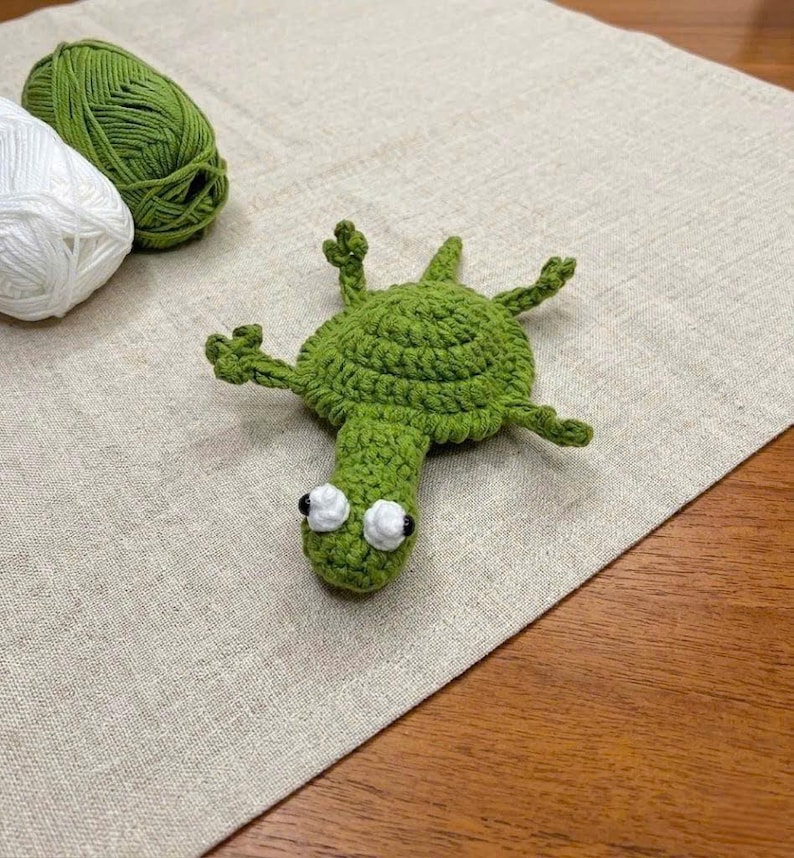 May include: A green crocheted turtle figure with a round shell, four small legs, and a head featuring two white eyes with black pupils. The turtle is positioned near a ball of green yarn and a ball of white yarn on a textured fabric surface.