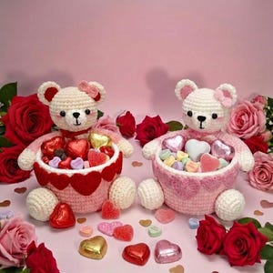 Puede incluir: Dos osos de peluche de ganchillo hechos a mano, cada uno sosteniendo una cesta de caramelos en forma de corazón. Los osos son blancos con detalles rosas y lazos. Las cestas son rosas y rojas, rodeadas de rosas rojas y caramelos en forma de corazón.