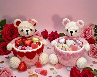 May include: Two handmade crochet teddy bears, each holding a basket of heart-shaped candies. The bears are white with pink accents and bows. The baskets are pink and red, surrounded by red roses and scattered heart-shaped candies on a pink surface.