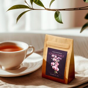 Może przedstawiać: Filiżanka herbaty i torebka herbaty rumiankowej leżą na stole. Torebka herbaty ma fioletową etykietę ze słowem "Chamomile" i kwiatowym wzorem. Filiżanka jest biała i stoi na białym spodku. W tle znajduje się zieleń.