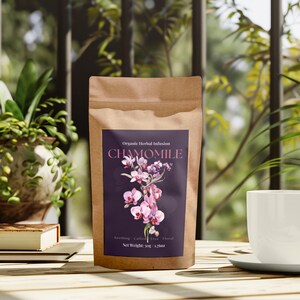 Może przedstawiać: Brązowa papierowa torebka z herbatą rumiankową z fioletową etykietą z kwiatowymi grafikami i napisem "CHAMOMILE". Biała filiżanka i spodek stoją na drewnianym stole, obok rośliny doniczkowej i książki.