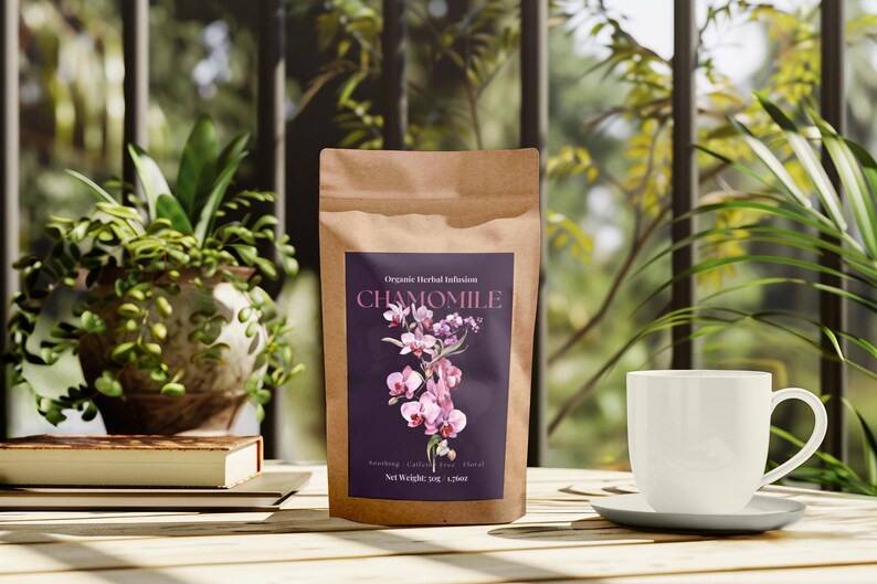 Może przedstawiać: Brązowa papierowa torebka z herbatą rumiankową z fioletową etykietą z kwiatowymi grafikami i napisem "CHAMOMILE". Biała filiżanka i spodek stoją na drewnianym stole, obok rośliny doniczkowej i książki.