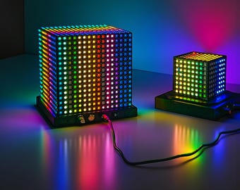 Puede incluir: Dos lámparas LED con forma de cubo que muestran un arcoíris de colores. La lámpara más grande es rectangular, y la más pequeña es cuadrada. Ambas lámparas están sobre bases negras y están conectadas por cables negros. Las luces crean una exhibición vibrante y colorida.