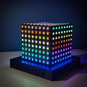 Puede incluir: Una pantalla de luz LED en forma de cubo con un arcoíris de colores. El cubo está sentado sobre una base negra con la palabra "Cubeworld" impresa en ella.