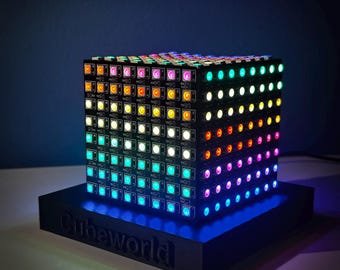 Puede incluir: Una pantalla de luz LED en forma de cubo con un arcoíris de colores. El cubo está sentado sobre una base negra con la palabra "Cubeworld" impresa en ella.