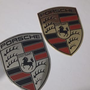 Op de afbeelding: Twee Porsche embleem badges. Eén is zilver met zwarte, rode en witte details, en de andere is goudkleurig met zwarte, rode en bruine details. Beide hebben de Porsche naam en de tekst Stuttgart.
