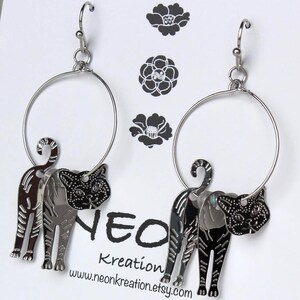 May include: Silver hoop earrings with a black cat charm hanging from each hoop. The cat charms are detailed with a striped pattern and have a black and white face.