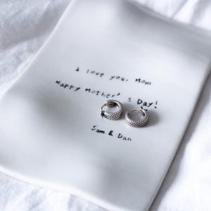 Puede incluir: Un plato de cerámica blanco con el mensaje escrito a mano "I love you, Mom. Happy Mother's Day! Sam & Dan." Dos pendientes de aro plateados con un diseño texturizado descansan sobre el plato.