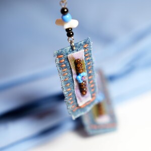May include: Close-up of a handmade earring with a denim rectangle base, adorned with colorful beads and stitching. The earring features a combination of blue, brown, and white beads, with a light blue background. The design is artistic and unique.