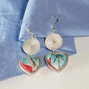 May include: A pair of silver earrings with a spiral design above heart-shaped pendants. The pendants feature embroidered designs in red, teal, and white. The earrings hang against a light blue fabric background.