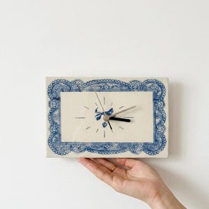 May include: Rectangular ceramic clock with a blue lace-like border and a central design featuring a bow and heart. The clock hands are black and silver. The clock is held by a hand.