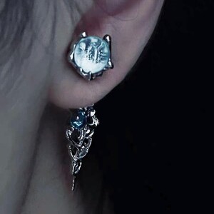 May include: Silver dangle earrings with a clear crystal stone and a smaller blue crystal stone. The earrings have a delicate, intricate design.
