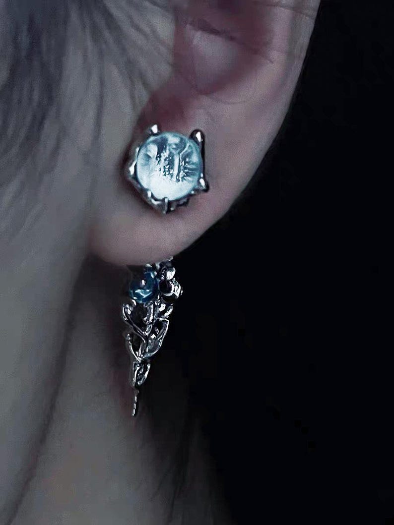 May include: Silver dangle earrings with a clear crystal stone and a smaller blue crystal stone. The earrings have a delicate, intricate design.