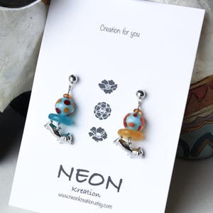 May include: A pair of silver earrings with blue and orange glass beads. The beads are decorated with white polka dots. The earrings are displayed on a white card with the text "Creation for you" and "NEON Kreation".