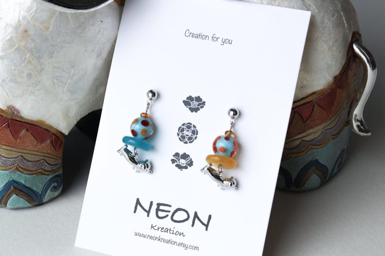May include: A pair of silver earrings with blue and orange glass beads. The beads are decorated with white polka dots. The earrings are displayed on a white card with the text "Creation for you" and "NEON Kreation".