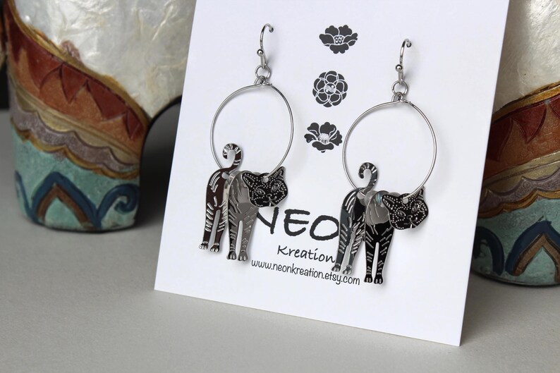 May include: Silver hoop earrings with a black cat charm hanging from each hoop. The cat charms are detailed with a striped pattern and have a black and white face.