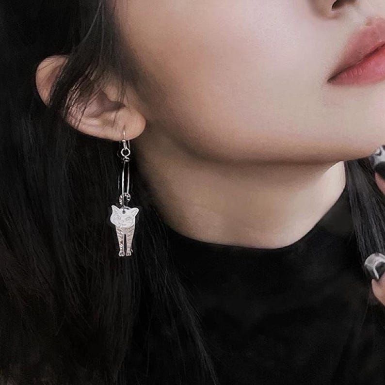 May include: Silver cat dangle earrings with a hoop design. The cat is facing forward with a white face and black eyes.