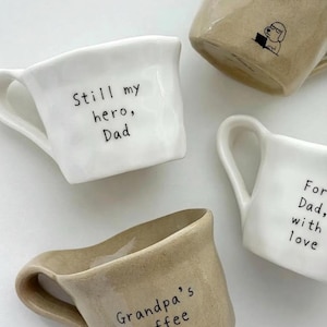 May include: Four ceramic mugs in white and beige. Two white mugs have the phrases "Still my hero, Dad" and "For Dad, with love." A beige mug says "Grandpa's coffee." Another beige mug has a dog reading a book.