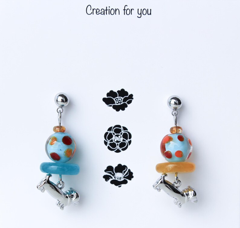 May include: A pair of earrings featuring a blue and orange glass bead with white polka dots, a silver dachshund charm, and a silver post.