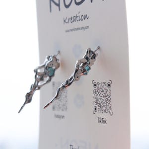 May include: A pair of silver earrings with a unique, organic design. Each earring features a delicate, twisted branch-like shape with a small, light blue gemstone at the end. The earrings are displayed on a white card with the text "Kreaton" and a QR code.