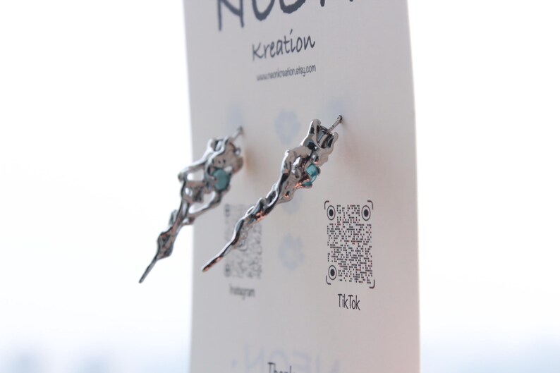May include: A pair of silver earrings with a unique, organic design. Each earring features a delicate, twisted branch-like shape with a small, light blue gemstone at the end. The earrings are displayed on a white card with the text "Kreaton" and a QR code.