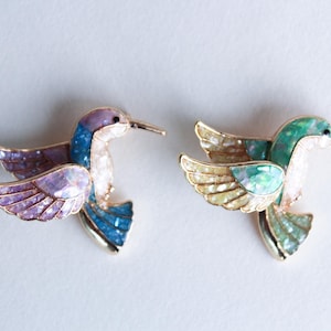 Puede incluir: Dos broches de colibrí con alas y cuerpos iridiscentes. Uno es azul, verde y morado, el otro es verde, amarillo y blanco.