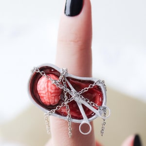 May include: A silver-toned ring featuring a miniature brain and red interior, secured with chains. The ring also includes small silver scissors. The ring is worn on a finger with a black nail polish.