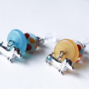 May include: A pair of dangle earrings featuring a silver dachshund charm, a blue and white glass bead, and a yellow and red glass bead.