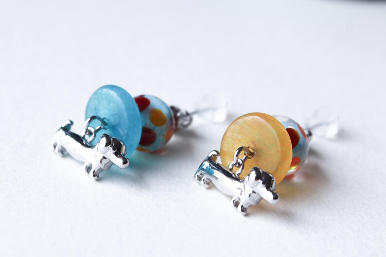 May include: A pair of dangle earrings featuring a silver dachshund charm, a blue and white glass bead, and a yellow and red glass bead.