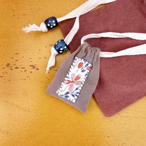 Può includere: Un piccolo sacchetto grigio con coulisse e un motivo floreale sul davanti. Il sacchetto ha un cordoncino bianco ed è impreziosito da due perline blu scuro con pois bianchi. Lo sfondo è di un caldo colore giallo-arancio.