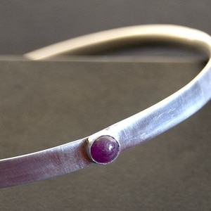 May include: A silver bangle bracelet with a single, round, purple gemstone set in the centre.