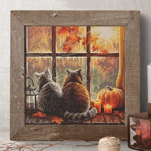 Peut inclure: Image de broderie au point de croix dans un cadre en bois rustique. L'image représente deux chats regardant par une fenêtre une scène d'automne avec une citrouille, des bougies et des feuilles d'automne. La palette de couleurs comprend des nuances de marron, d'orange et de gris.