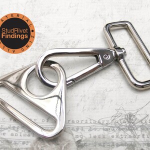 May include: Silver-toned metal swivel hook with a triangular D-ring and a rectangular D-ring. The hook is attached to the triangular D-ring. The text "StudRivet Findings" is printed on a small orange circle.
