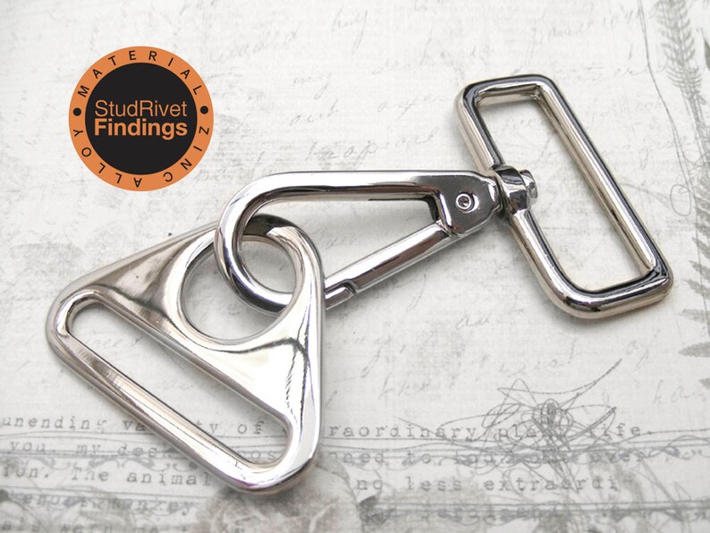 May include: Silver-toned metal swivel hook with a triangular D-ring and a rectangular D-ring. The hook is attached to the triangular D-ring. The text "StudRivet Findings" is printed on a small orange circle.